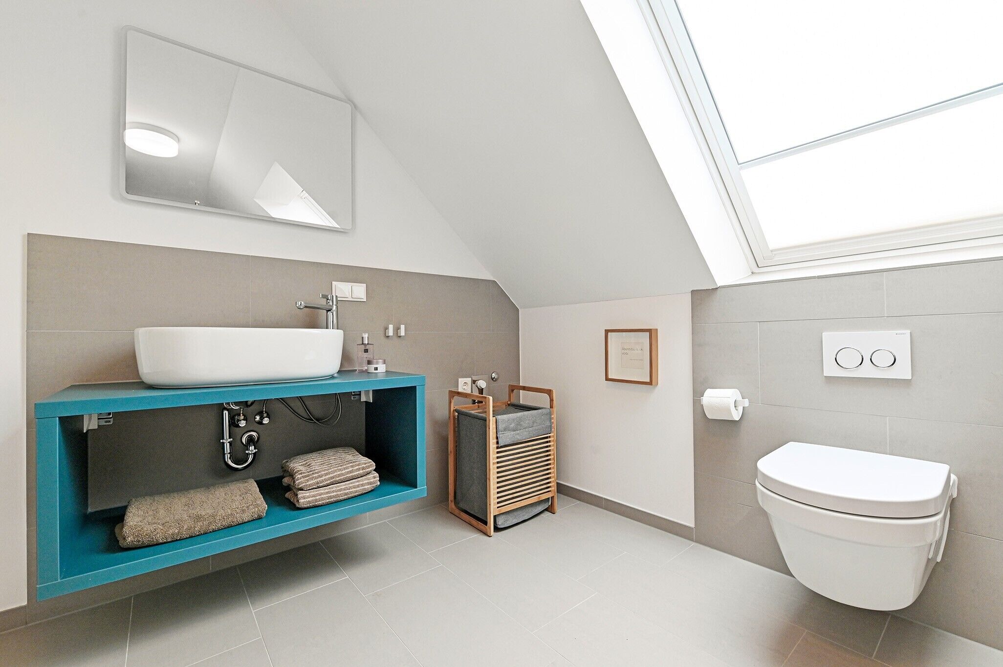 A modern bathroom with a skylight, a white sink, a blue vanity, and a white toilet.