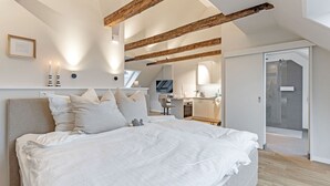 A modern bedroom with a large bed, wooden beams, a skylight, a kitchenette, and a sliding glass door to a balcony. - Achterdörn Een – Beautiful Loft for Relaxation (Fehmarn)