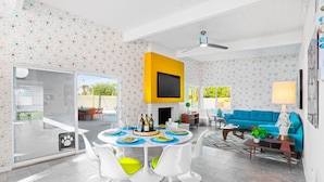 A modern living room with a dining area, a blue sofa, and a TV mounted on a yellow wall.