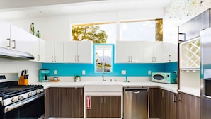 A modern kitchen with a stove, microwave, and dishwasher, featuring white and teal cabinetry and a window with a view of trees.