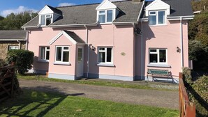 A pink two-story house with a chimney, a front door, and a small garden area with a bench. - 'Home From Home' 3 Bedroom Cottage in Nolton Haven (Haverfordwest)