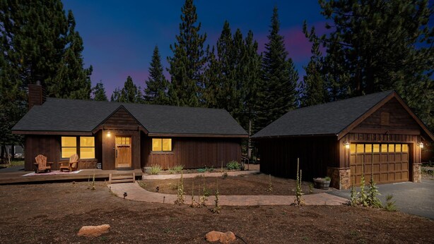 Une maison en bois à deux étages, avec un garage, une véranda à l’avant dotée de chaises et un jardin aménagé.