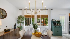 A dining area with a wooden table, chairs, a chandelier, a clock, a potted plant, and a view through the window.