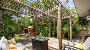 A covered patio with outdoor furniture, a play area, and lush greenery.