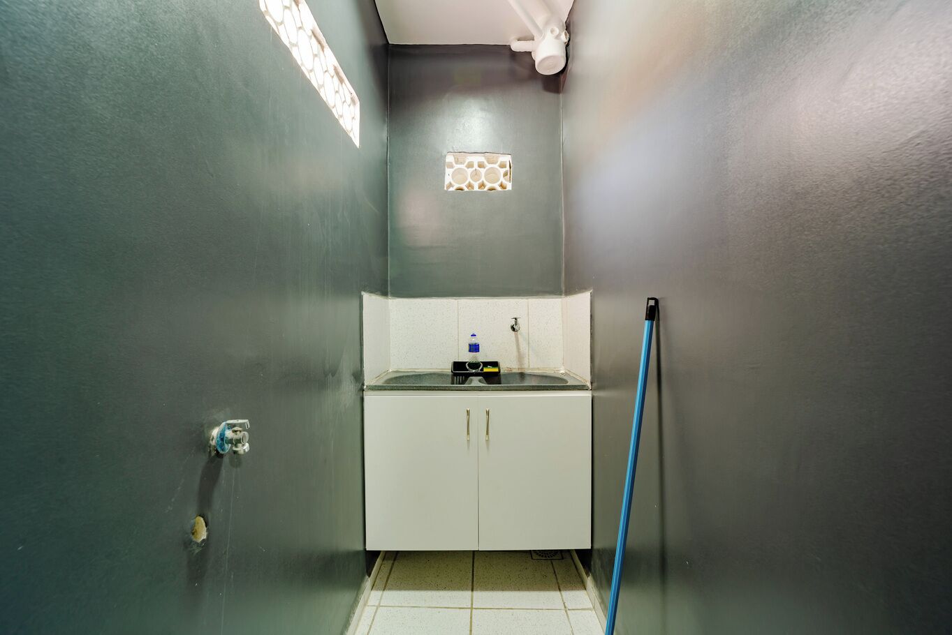 A small, enclosed bathroom with a sink, mirror, and a mop leaning against the wall.