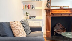A living room with a grey sofa, patterned cushions, a bookshelf with books, a fireplace, and a small table. - Victorian appartement in Marylebone (London)