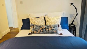A neatly made bed with a dark blue bedspread, two pillows with a floral pattern, and a blue pillow. A black floor lamp is on a small table beside the bed. - Victorian appartement in Marylebone (London)