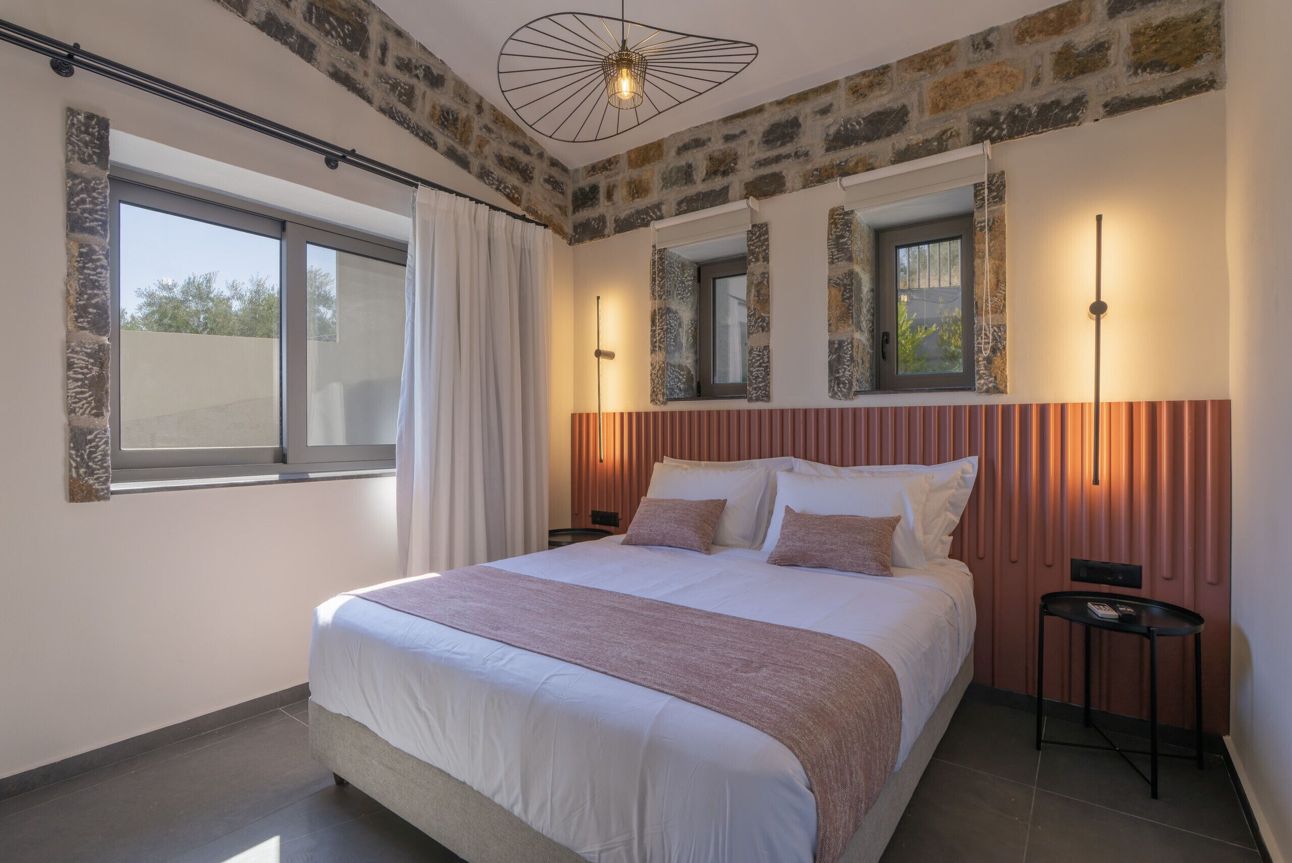 Un dormitorio con una cama grande, una ventana con cortinas, un muro de piedra y una lámpara moderna.