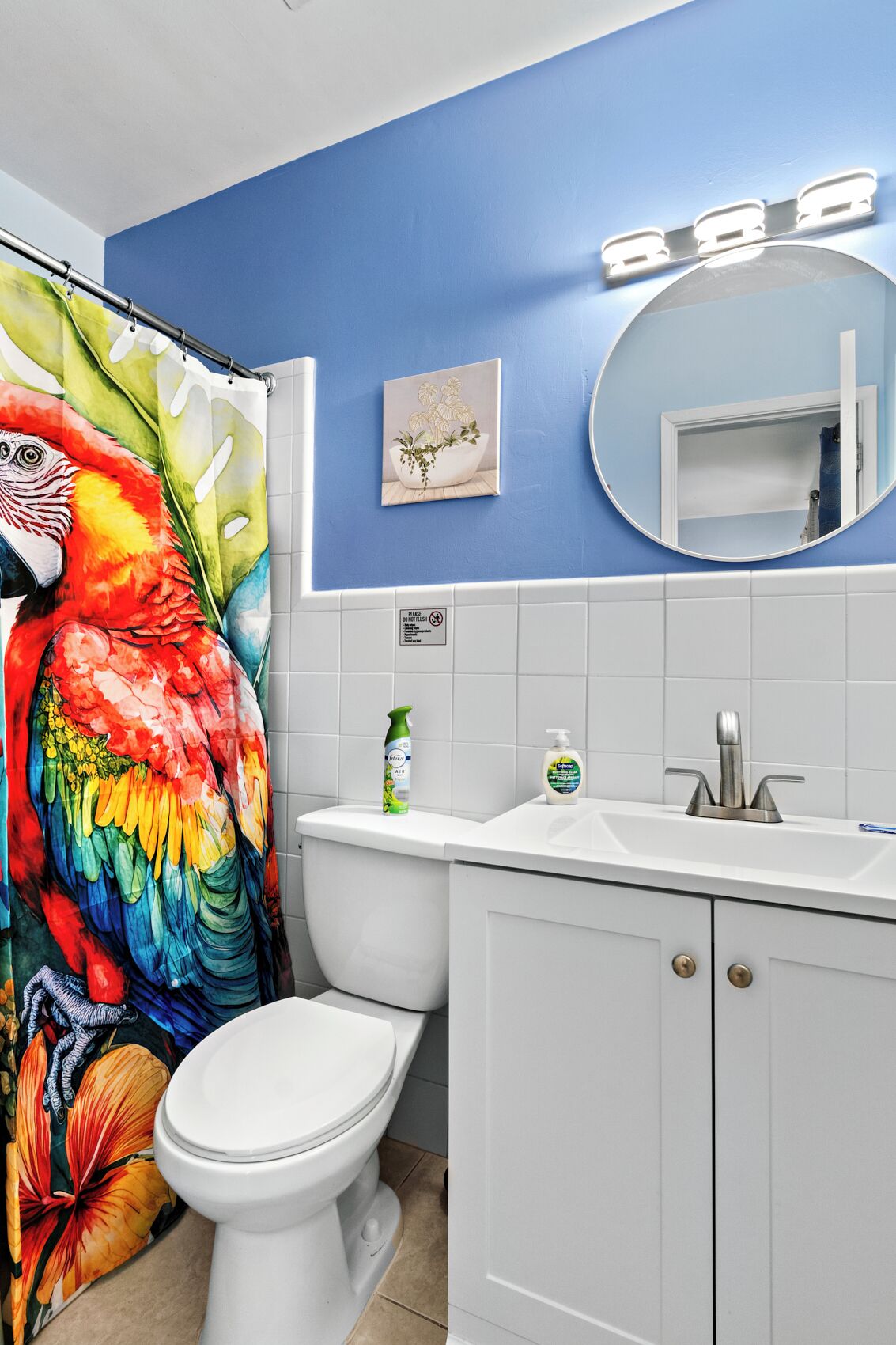 Une salle de bain avec un mur bleu, des toilettes blanches, un rideau de douche orné d’un motif coloré représentant un perroquet, un lavabo avec un miroir et des articles de toilette posés sur le comptoir.