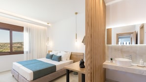 A modern bedroom with a large bed, a bedside table, a nightstand, a window with curtains, a bathroom with a sink and mirror, and a view of a desert landscape. - Villa Santa Irini 3 - With Heated Pool (Rethymno)