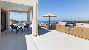 A rooftop terrace with a dining table and chairs, a grill station, and lounge chairs under umbrellas, overlooking a coastal view. - Villa Santa Irini 3 - With Heated Pool (Rethymno)