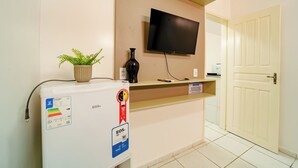 A hotel room with a minibar, a television mounted on the wall, a potted plant, and a door to the right. - Flat Para 2 Pessoas a Poucos Metros do Aeroporto (Fortaleza)