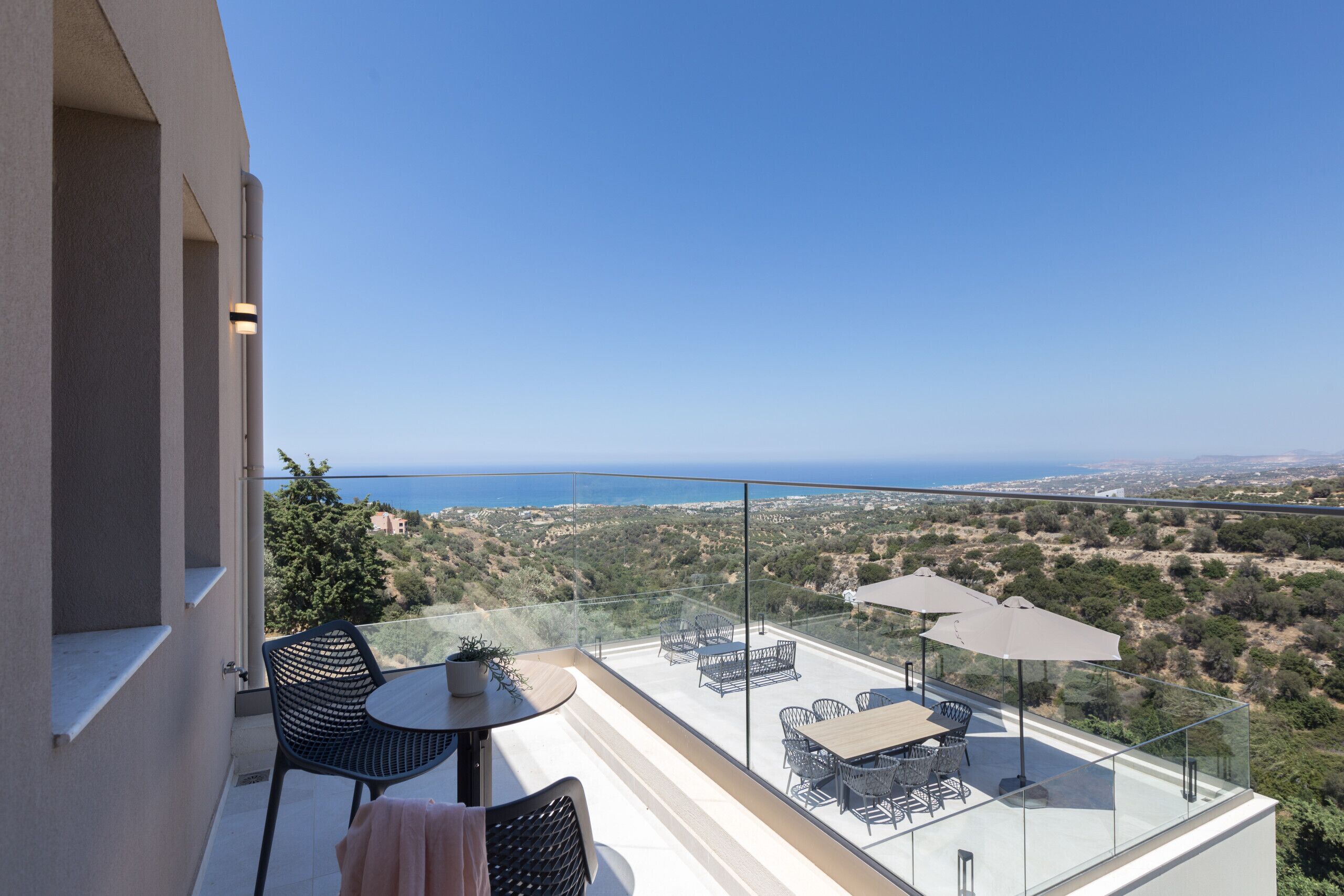 A balcony with a glass railing, outdoor furniture, and a view of the sea and landscape.