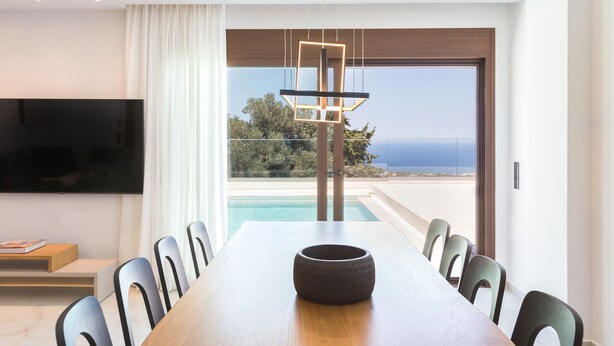 A modern dining area with a large table, chairs, a television, and a view of the ocean. - Villa Santa Irini 1 - With Heated Pool (Rethymno)