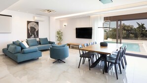 A modern living room with a sectional sofa, a dining table with chairs, a flat-screen TV, and a large window overlooking a swimming pool. - Villa Santa Irini 1 - With Heated Pool (Rethymno)