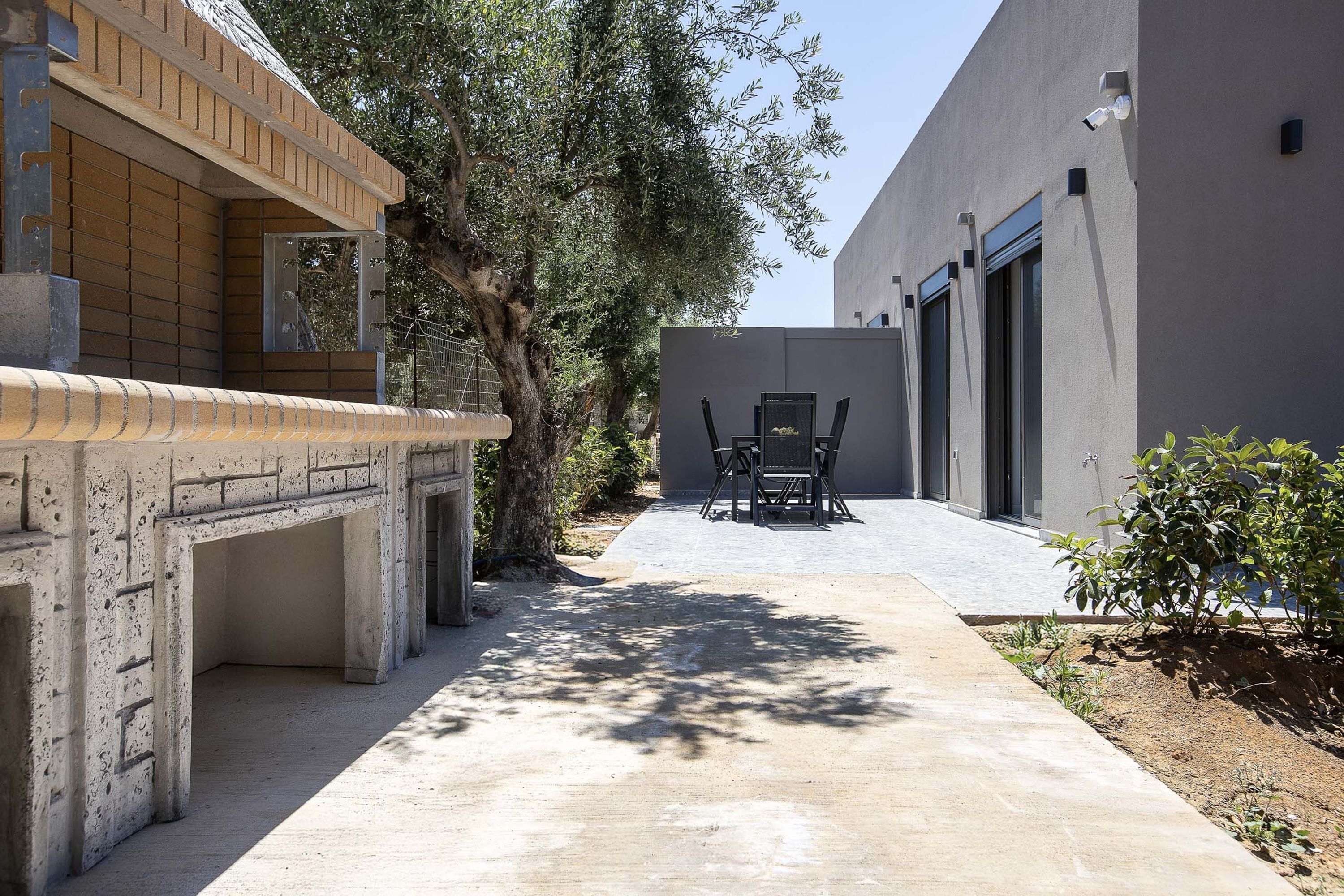 Une cour avec un chemin en béton, un banc en bois et une table avec des chaises. On y trouve un arbre et un bâtiment doté de grandes fenêtres.