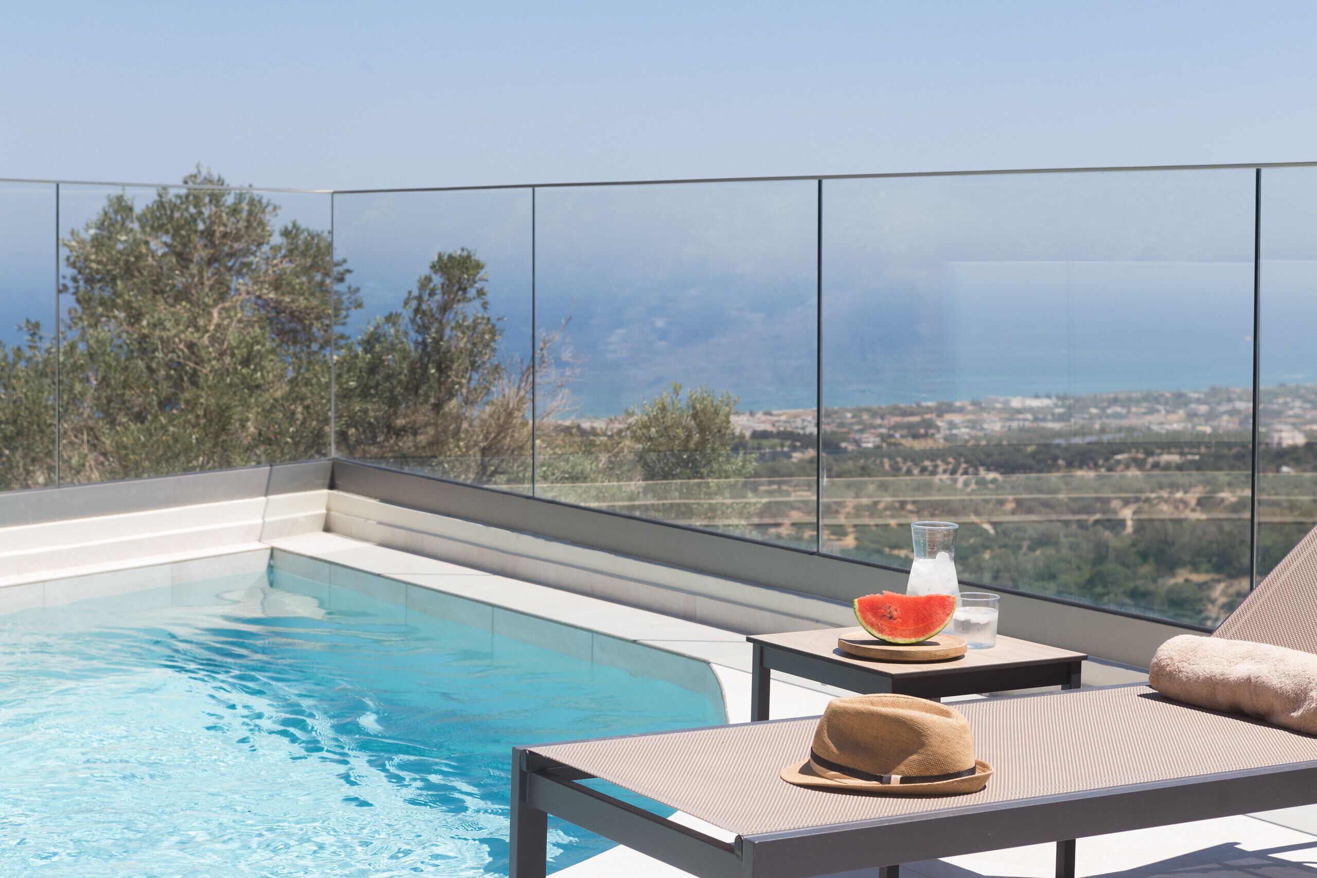 A rooftop pool with clear blue water, a glass of water, a slice of watermelon, and a hat on a lounge chair.