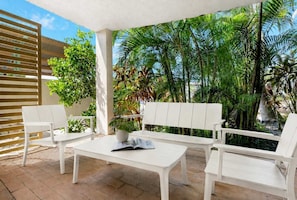 A covered patio area with white furniture, including a bench and chairs, surrounded by lush greenery and palm trees.