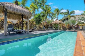 A swimming pool with lounge chairs and thatched umbrellas, surrounded by palm trees and tropical plants.
