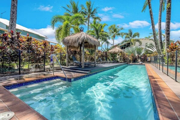 Ein Swimmingpool mit einem strohgedeckten Gebäude, umgeben von Palmen und tropischer Vegetation.
