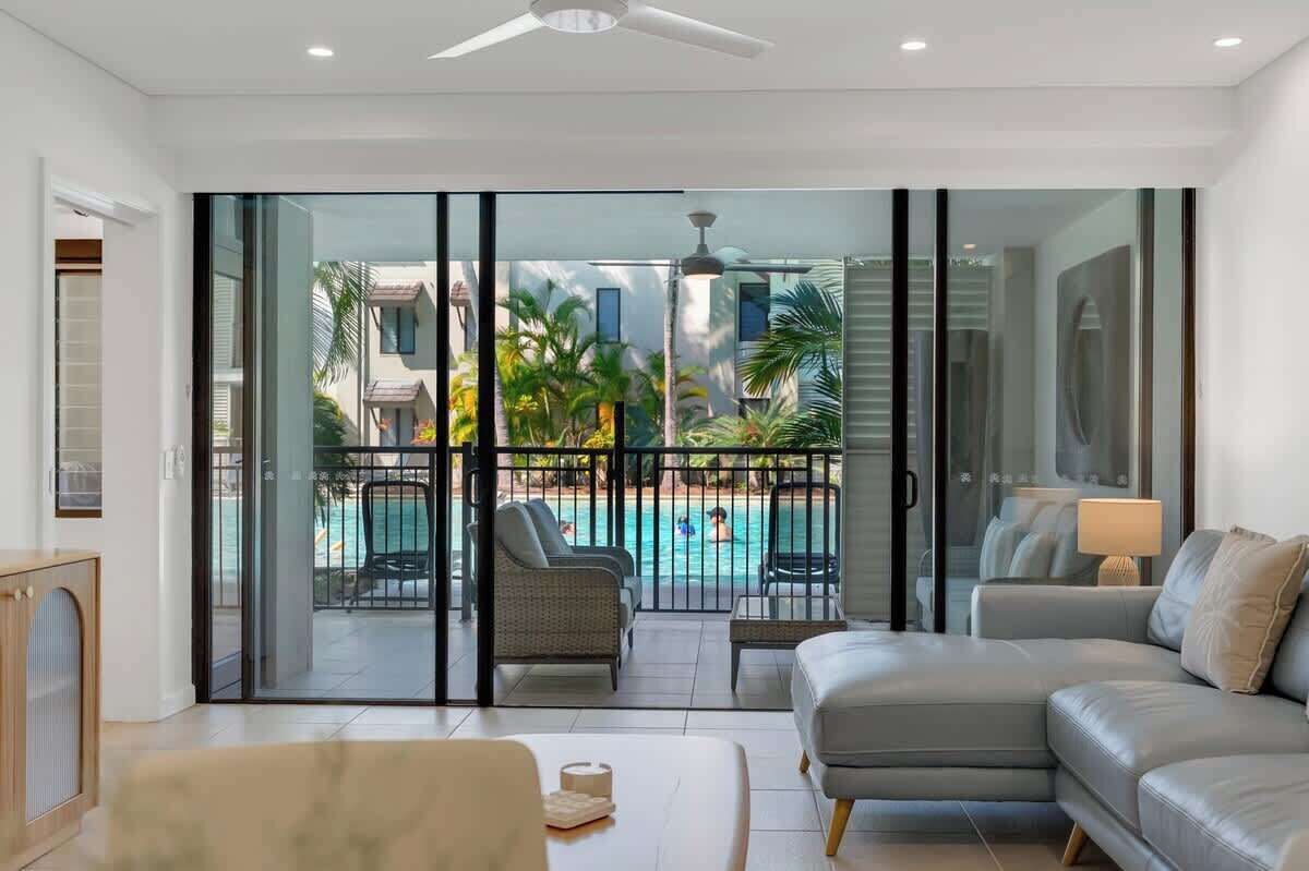 A modern living room with a sofa, a coffee table, and a view of a swimming pool through large glass doors.