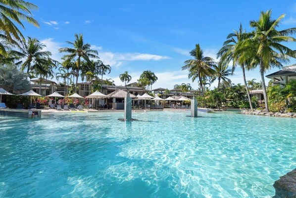 A clear blue pool surrounded by palm trees and beach umbrellas, with resort buildings in the background.
