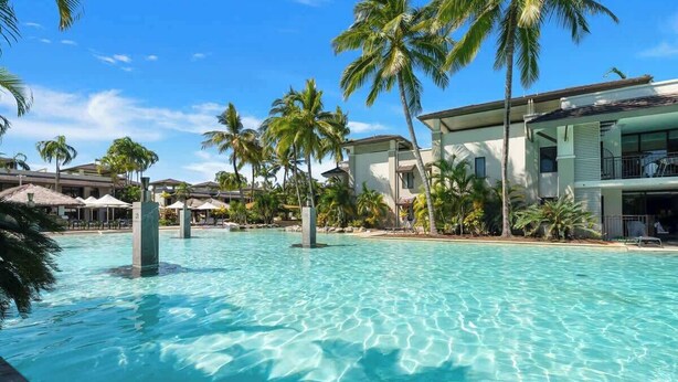 Une piscine d’un complexe hôtelier, avec de l’eau claire, des palmiers et les bâtiments du complexe.