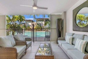 Un balcone con mobili in vimini, un tavolino da caffè in vetro e vista su una piscina e palme.