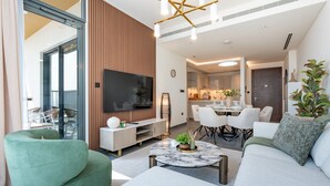 A modern living room with a sofa, a coffee table, and a TV. There is a dining area with chairs and a table. - Skyline Views 2BR, Steps from a Park, Minutes Drive to Meydan Golf Club (Dubai)