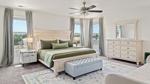 Un dormitorio con cama, mesitas de noche, cómoda, ventilador de techo y ventanas amplias.