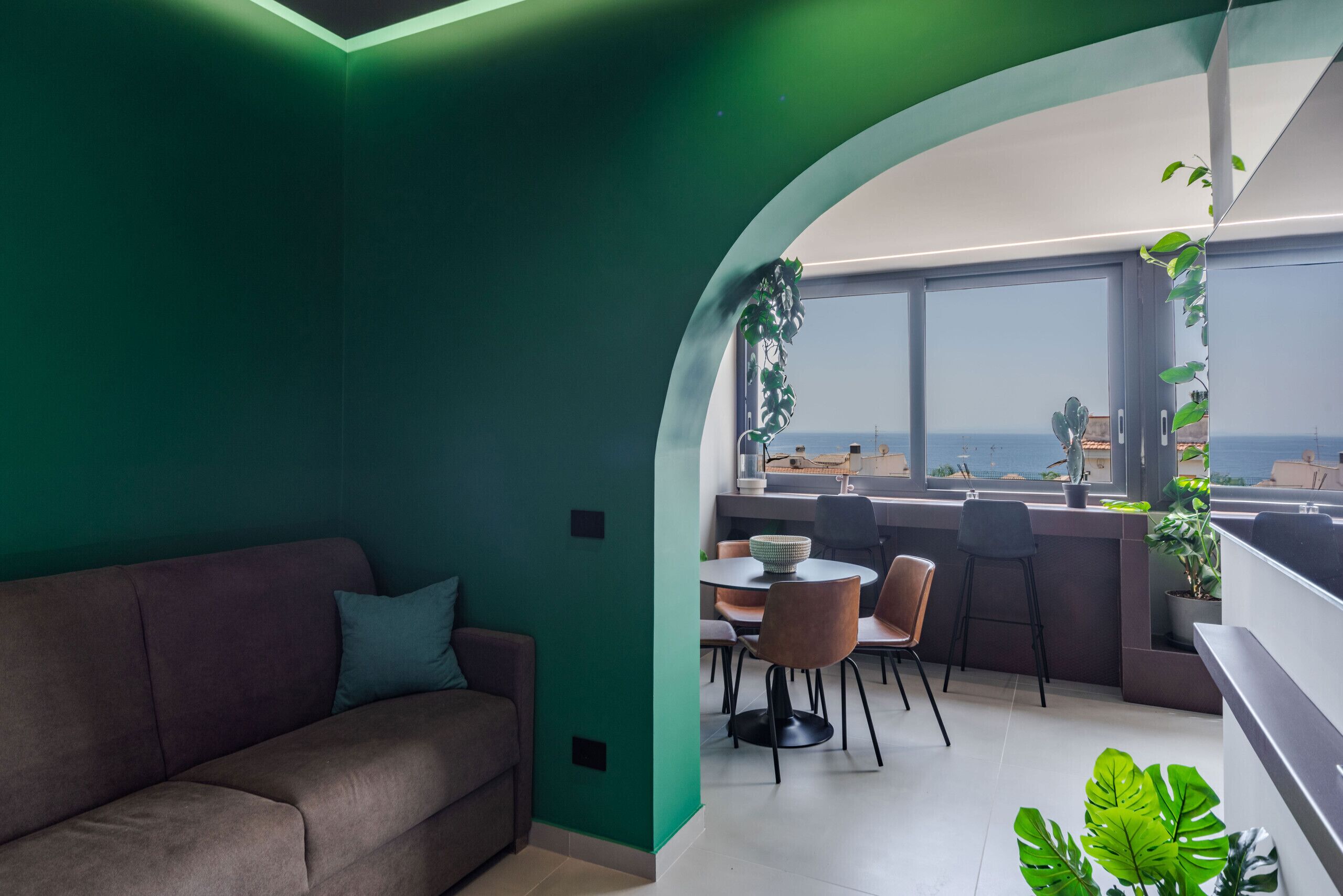 Ein modernes Wohnzimmer mit grüner Akzentwand, einem braunen Sofa, einem Essbereich mit Stühlen und Tisch sowie einem Blick auf die Stadt durch große Fenster.