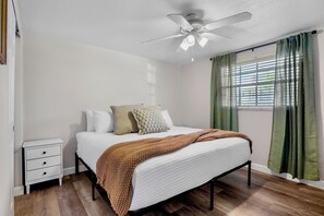 Un dormitorio con cama, mesita de noche, ventilador de techo y ventana con cortinas.