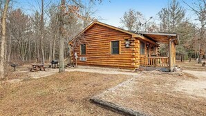 A log cabin with a porch, surrounded by trees and a picnic table.
