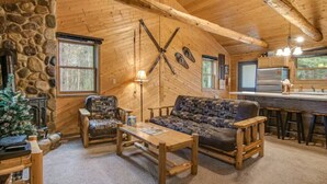 A cozy cabin interior with a stone fireplace, wooden furniture, and a kitchen area.