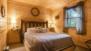 A wooden cabin room with a bed, two bedside lamps, a window with curtains, and a wall clock.