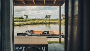 Une terrasse en bois avec une table et des chaises, donnant sur une rivière paisible où naviguent des bateaux.