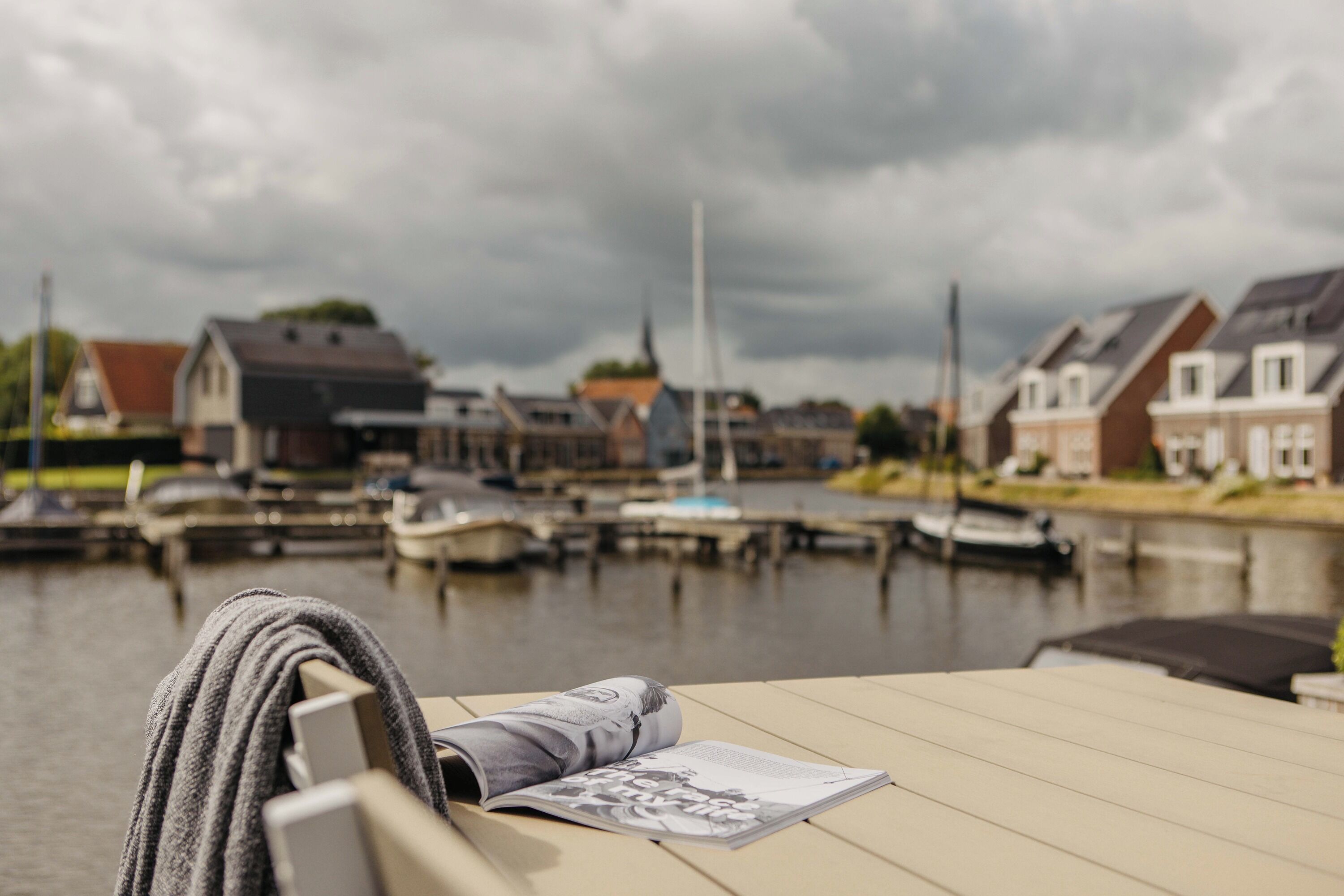 A view of a riverside area with boats docked, houses along the water, and a table with a magazine and a towel.