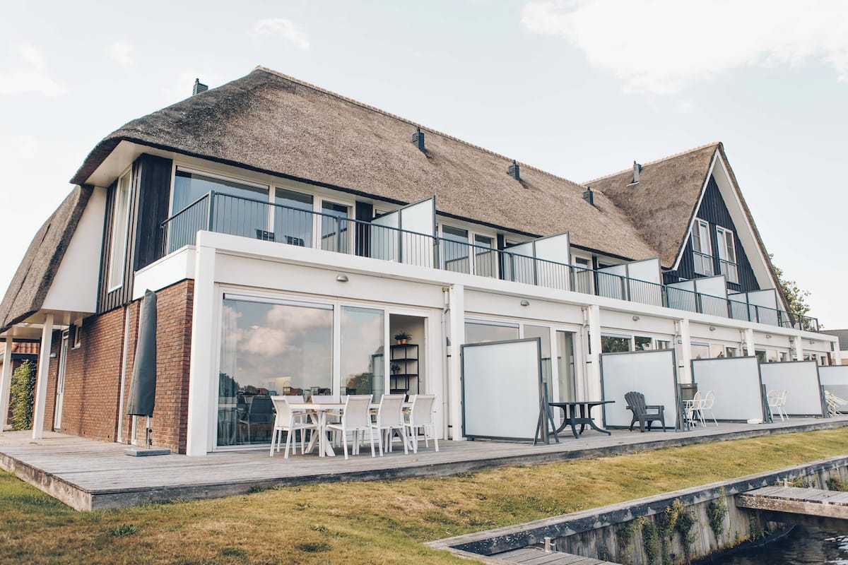A modern house with a large deck, a thatched roof, and a view of a canal.