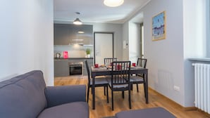 Moderni olohuone, jossa on sohva, ruokapöytä ja tuoleja. Taustalla näkyy keittiö, jossa on nykyaikaiset kodinkoneet ja kahvinkeitin.