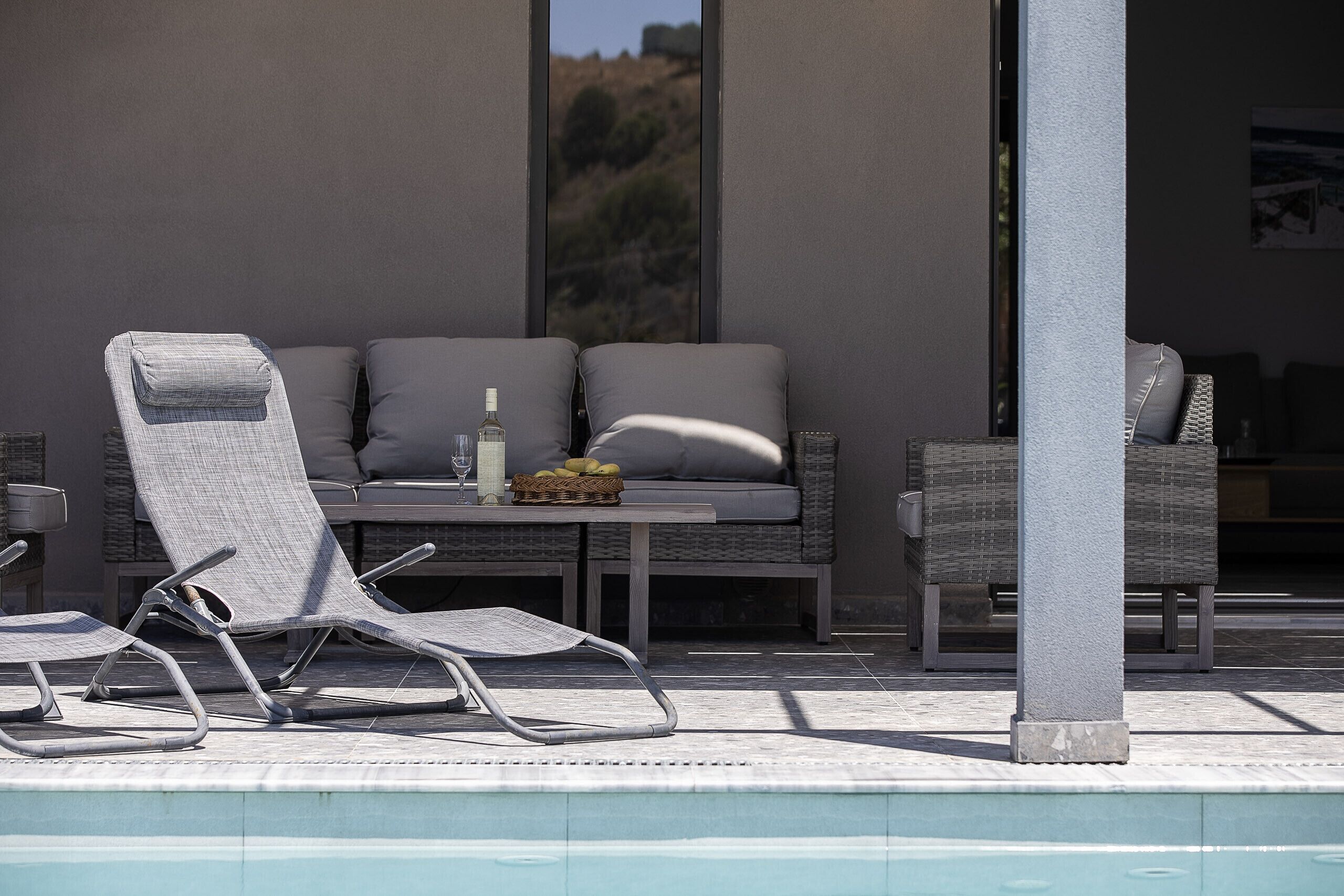 Un espace aménagé au bord de la piscine, avec des chaises longues, un canapé en osier et une table sur laquelle se trouvent une bouteille et un panier de fruits.