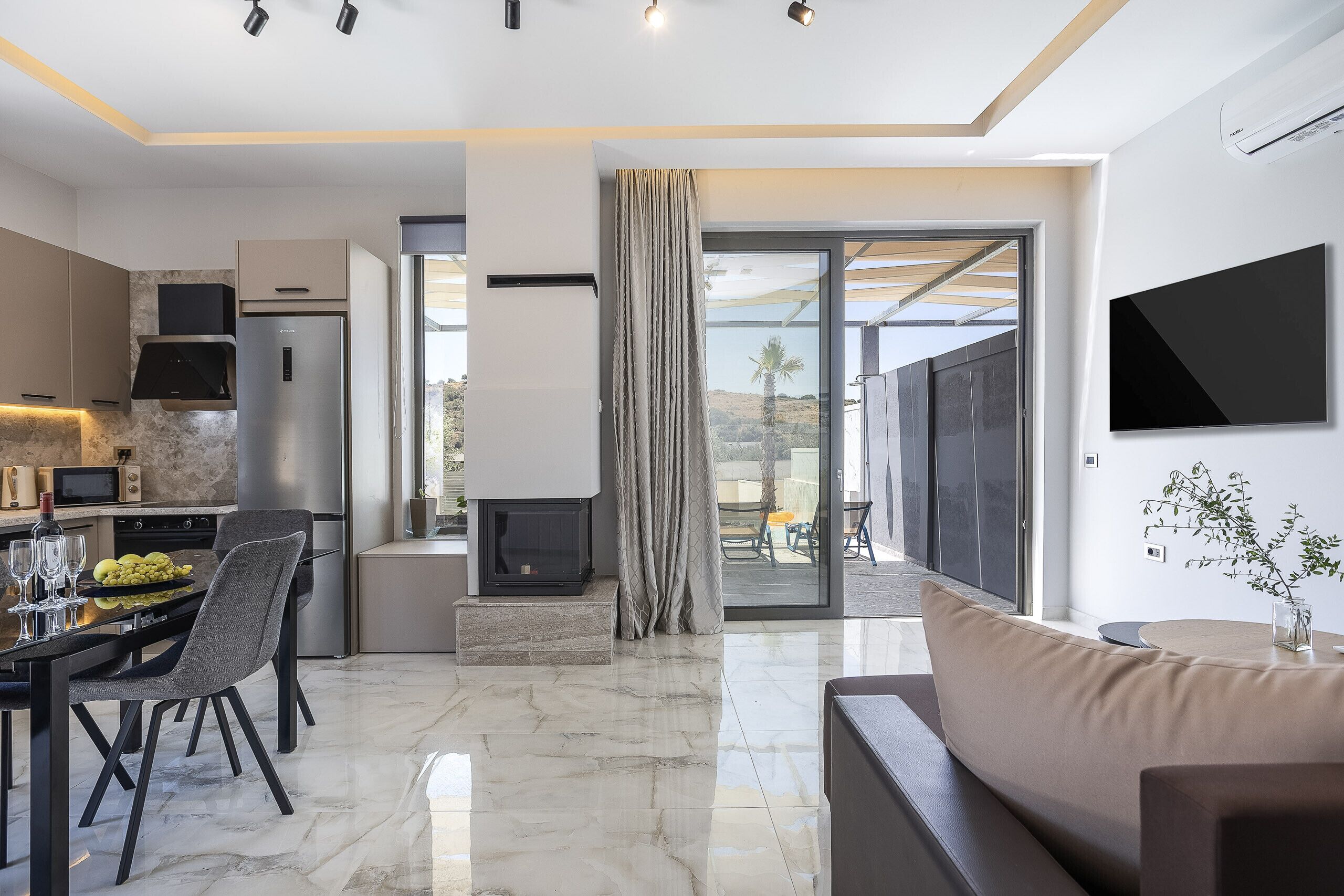 A modern kitchen with a dining area, a living room with a sofa, and a large window offering a view of the outdoors.