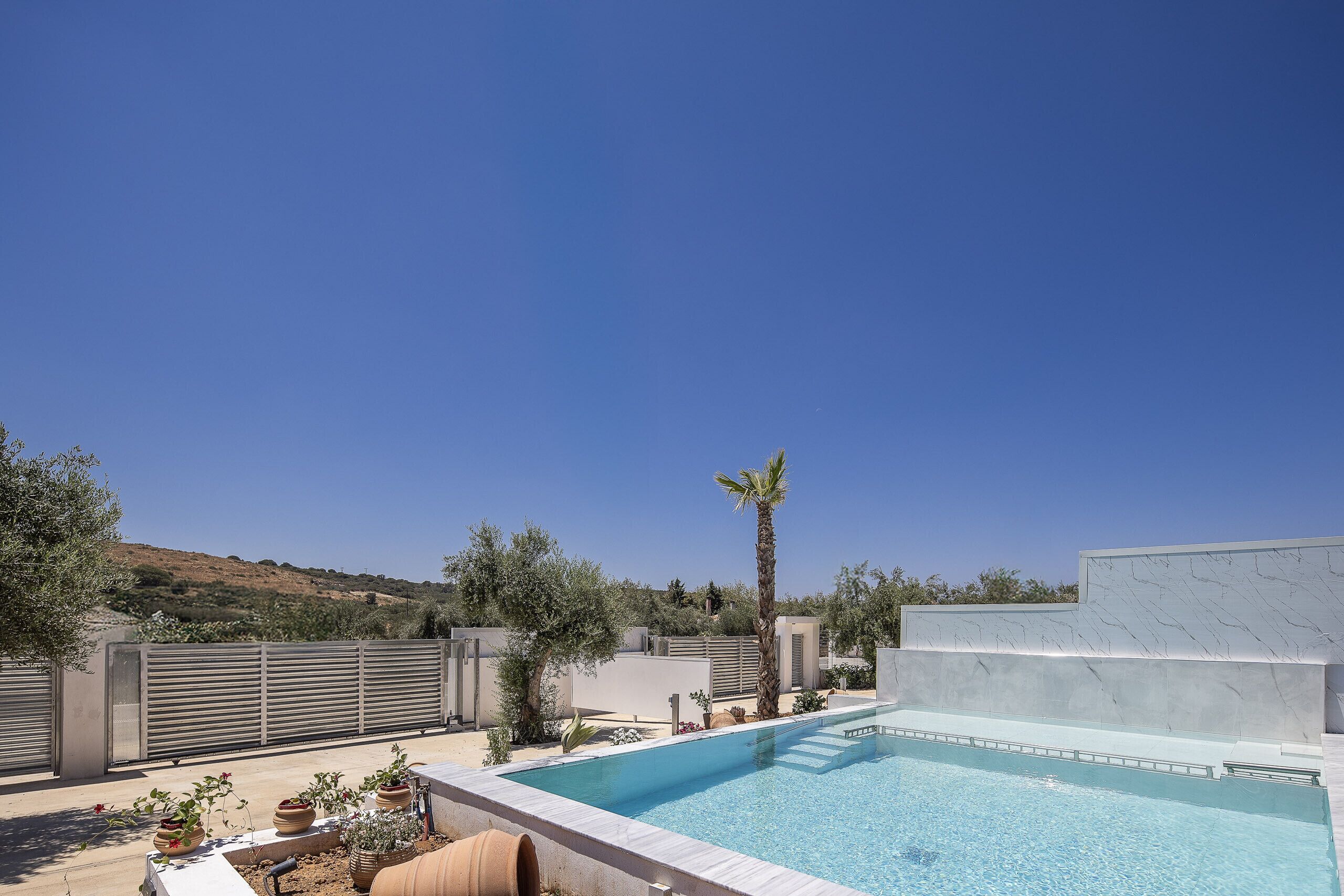A swimming pool with a clear blue sky, surrounded by a modern building with a marble facade, and a hilly landscape in the background.