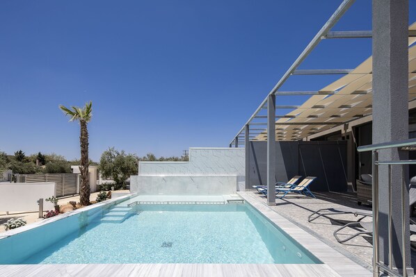 A swimming pool with a clear blue water, surrounded by a white tiled deck, and a pergola structure.