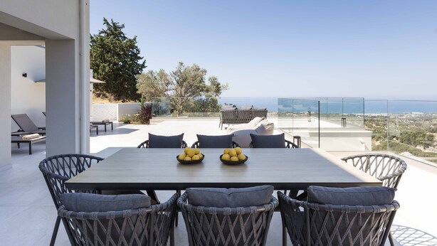 A modern outdoor dining area with a table and chairs, a view of the sea, and a clear sky. - Santa Irini Villas - With 3 Heated Pools (Rethymno)