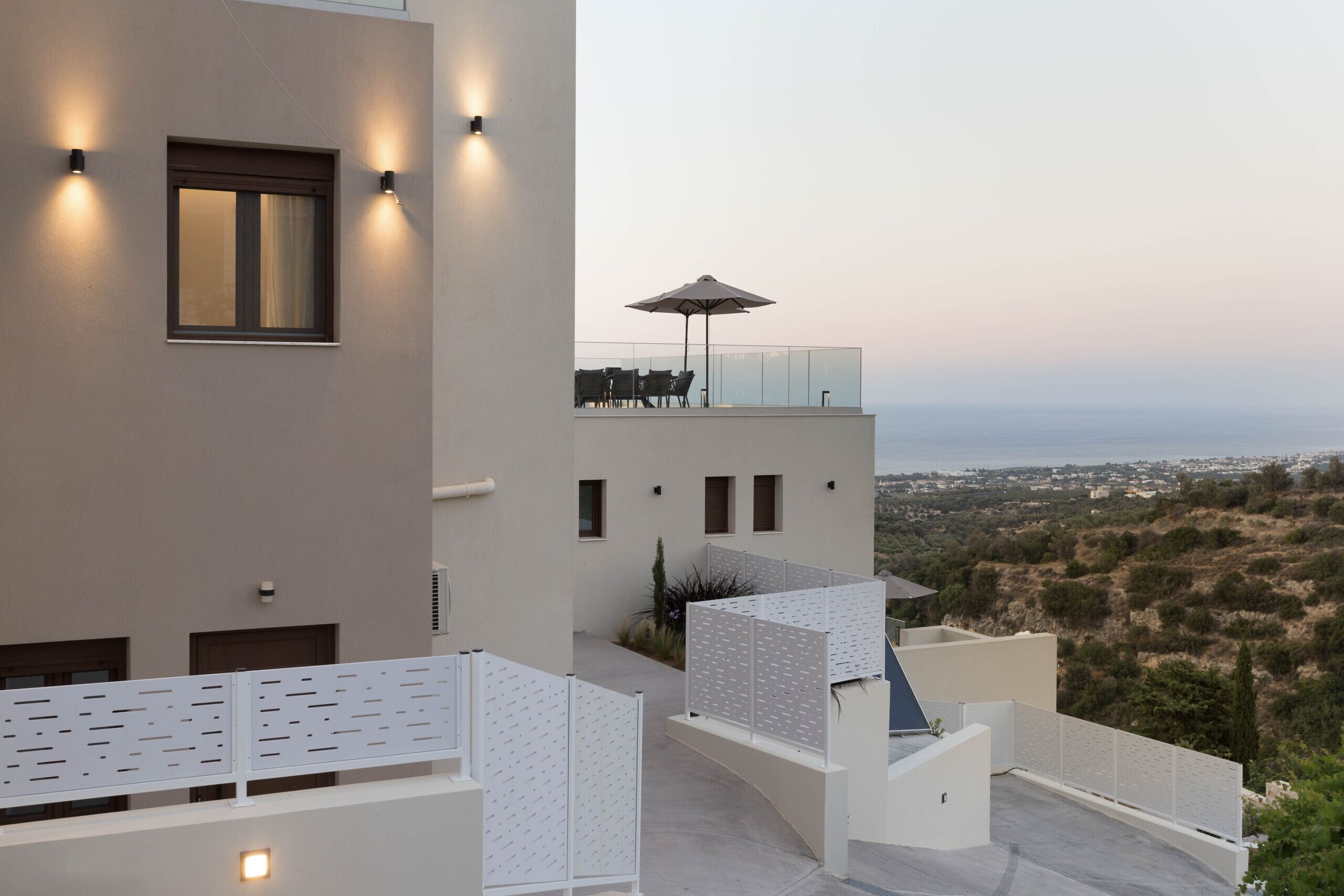 A modern building with a rooftop terrace featuring a glass balustrade, outdoor seating, and a view of the landscape.