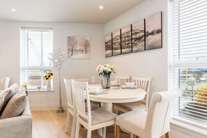 A dining area with a round table, white chairs, a vase with flowers, and a cityscape painting on the wall.