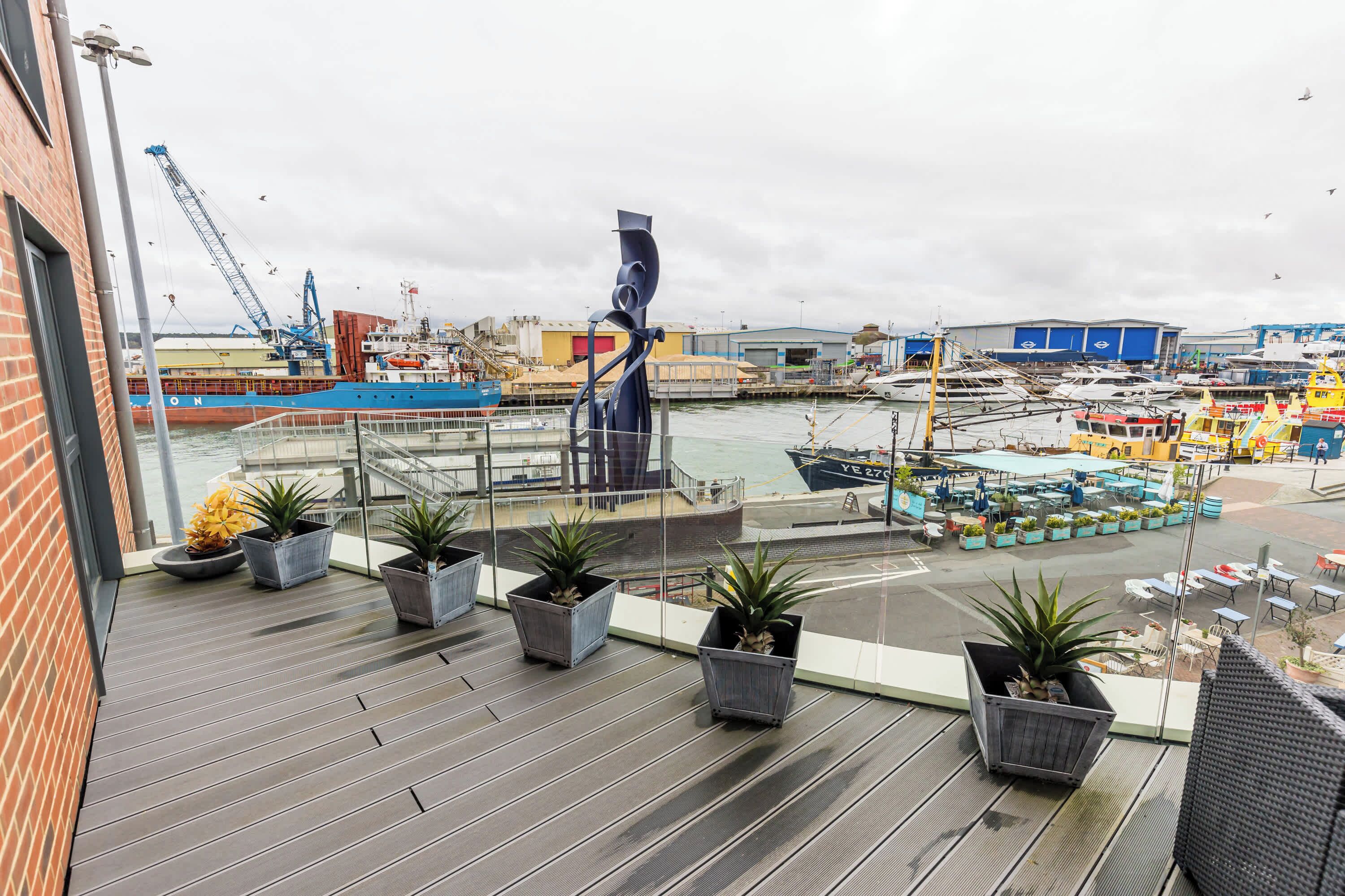 A balcony with potted plants, overlooking a marina with boats and a crane.