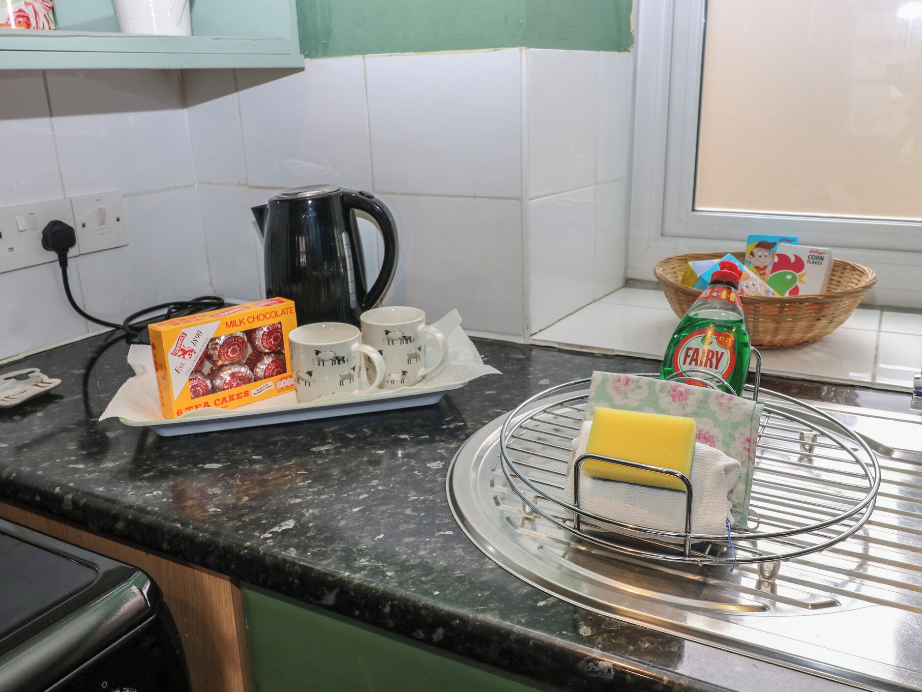 A kitchen counter with a box of milk chocolate cakes, two mugs, a kettle, a basket with cereal boxes, and a sponge on a drying rack.