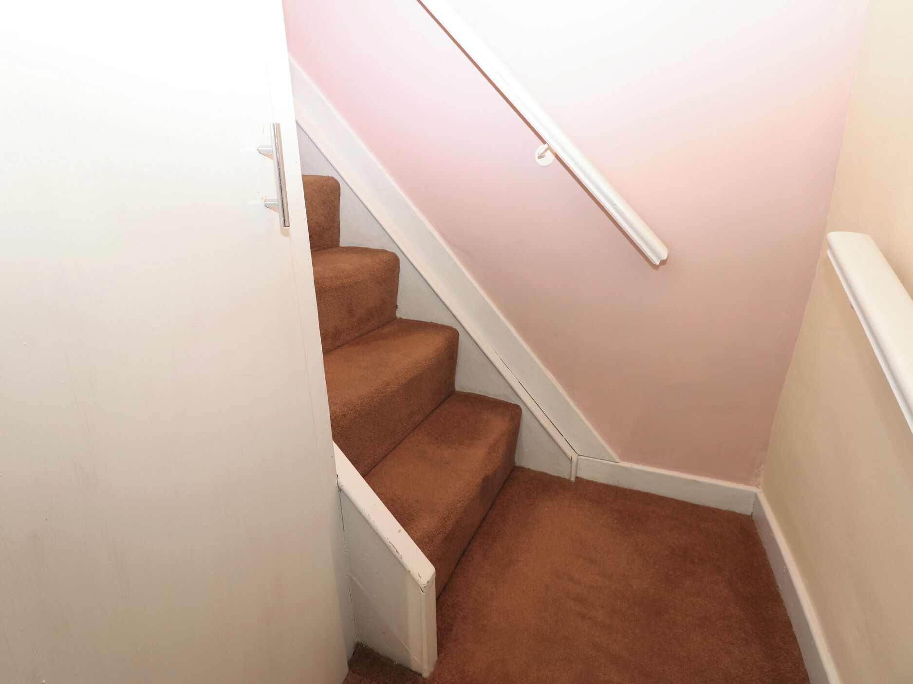 A staircase with carpeted steps and a white handrail, leading to a room with a pink wall.