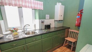 Une cuisine avec des meubles de couleur verte, un évier, une fenêtre avec un rideau rose, un radiateur blanc et un extincteur.